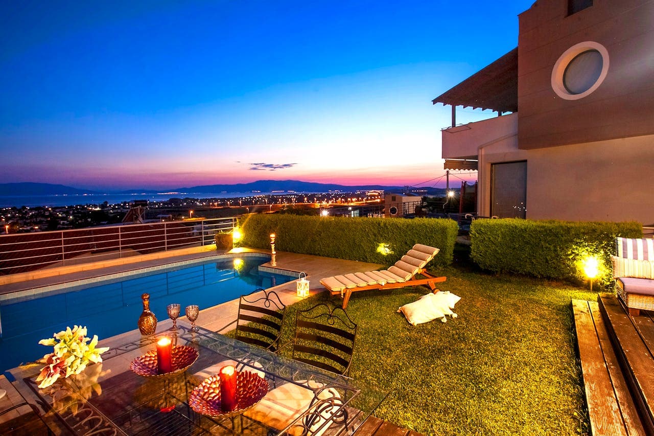 Exterior shot of a backyard with a pool, overlooking a city. The sun has set and there are lights on in the yard