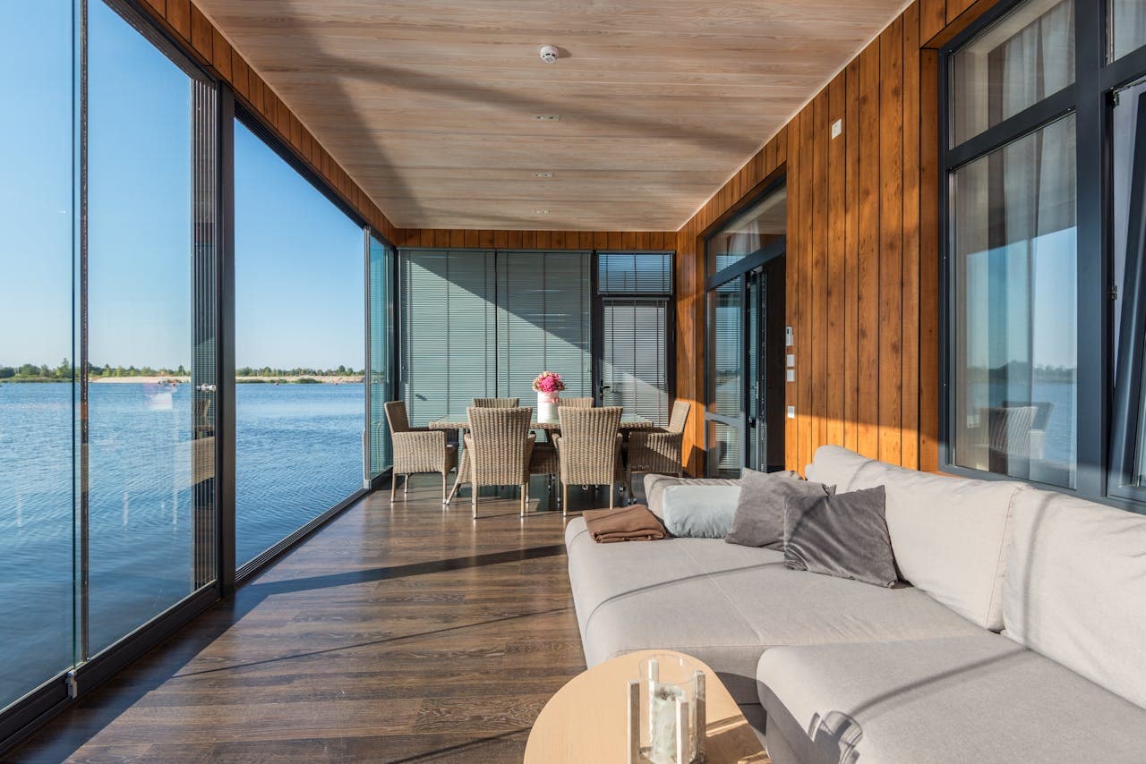 A green room with wooden paneling, couches, and a wicker-style table looks in front of a body of water