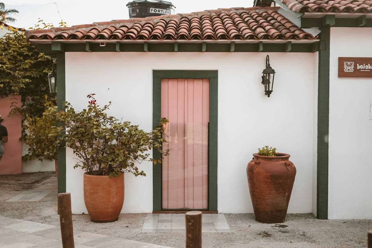 Close-up shot of a door leading into a small stucco cottage. There are two large pots holding plants on either side of the glass door